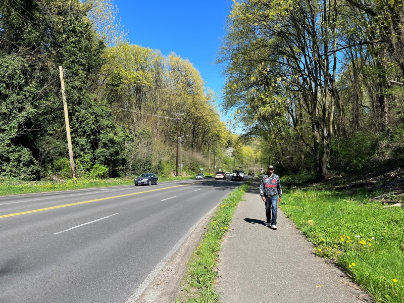 Persona que camina por el sendero existente a lo largo de Highland Park Way SW en un día soleado. A la izquierda del sendero hay una zona de protección paisajística con pasto que separa el sendero de la carretera. Junto a la zona de protección paisajística hay dos carriles de circulación en dirección norte por donde circulan dos vehículos uno al lado del otro. Más allá de eso, hay dos carriles de circulación hacia el sur con dos vehículos que circulan hacia el sur en el carril exterior.