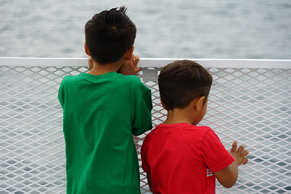 Two young children stand at the rail of the boat and look at the water.