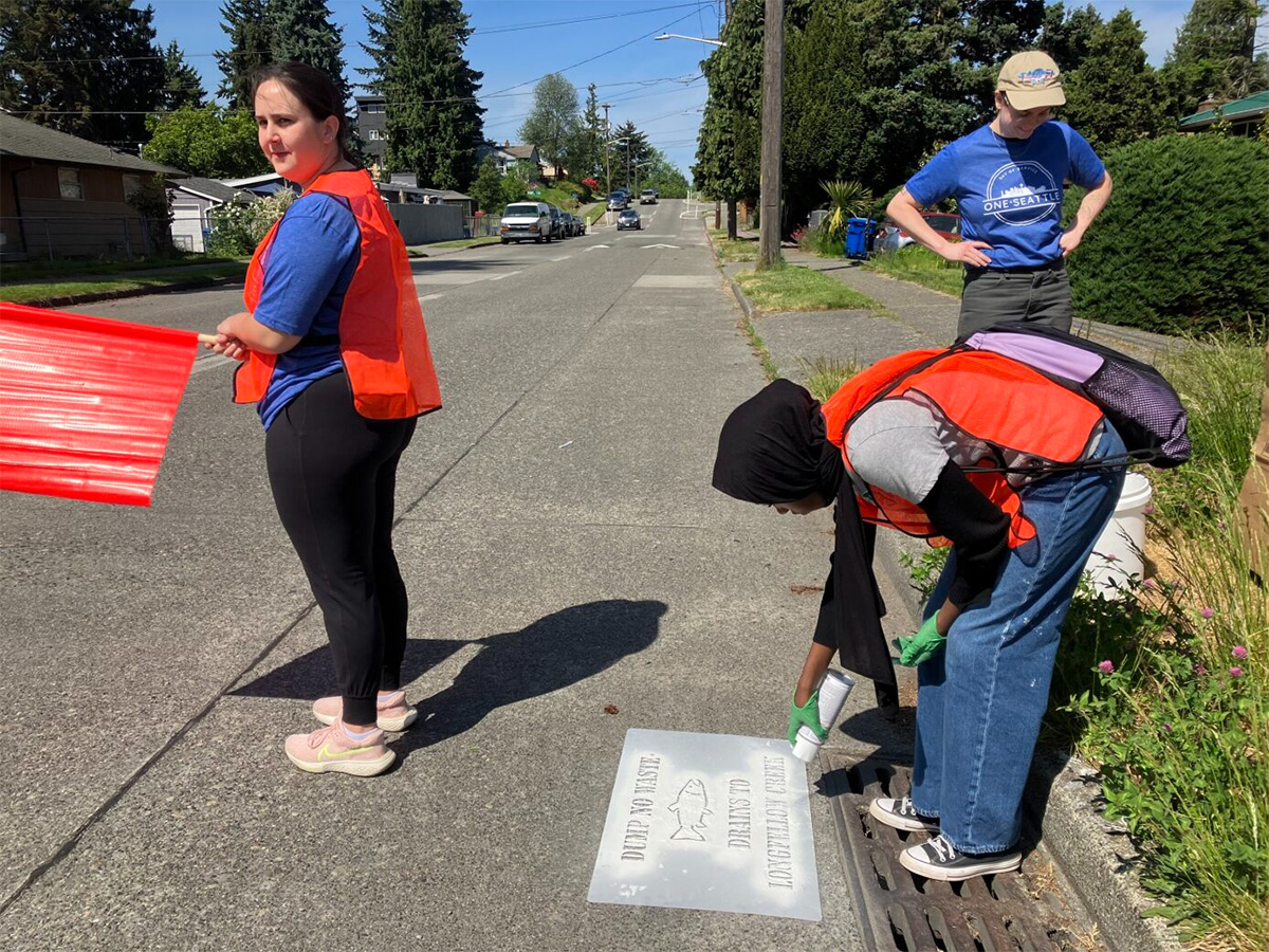 A flagger in an orange safety vest stands near another worker in an orange vest as they spray white paint on a stencil near a storm drain in the street.