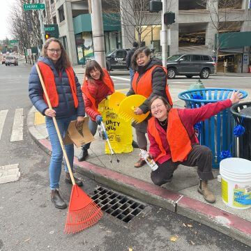 four people in orange safety vests next to a storm drain with cleaning tools.