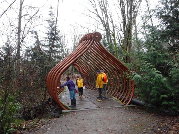 SPU staff examine the deck and railing of the Salmon Bone Bridge, a footbridge with overhead steel structures made to resemble fishbones surrounded by woods.