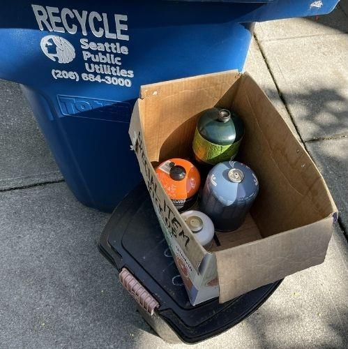 Propane canisters in a box near a recycling bin.