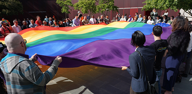 A giant pride flag is unfurled with a large group of people holding it around the edges. Pride flag raising, Seattle, 2018.