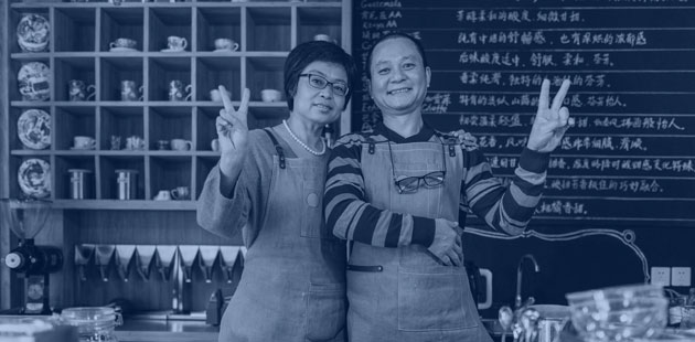 Two Asian American cafe owners stand behind the counter smiling and flashing "peace signs" with their fingers