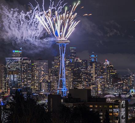 Fireworks launch from the top of the Space Needle at night with Seattle lit in green and blue