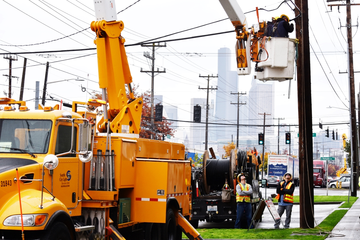Seattle City Light utility workers are performing maintenance on power lines. A bright orange utility truck with a crane and elevated bucket is positioned near the power lines, with one worker inside the bucket working on the lines. Another worker is on the ground, observing or assisting in the task. In the background, there’s an urban skyline with overcast skies. The presence of multiple utility poles and wires indicates this is an area with significant electrical infrastructure.