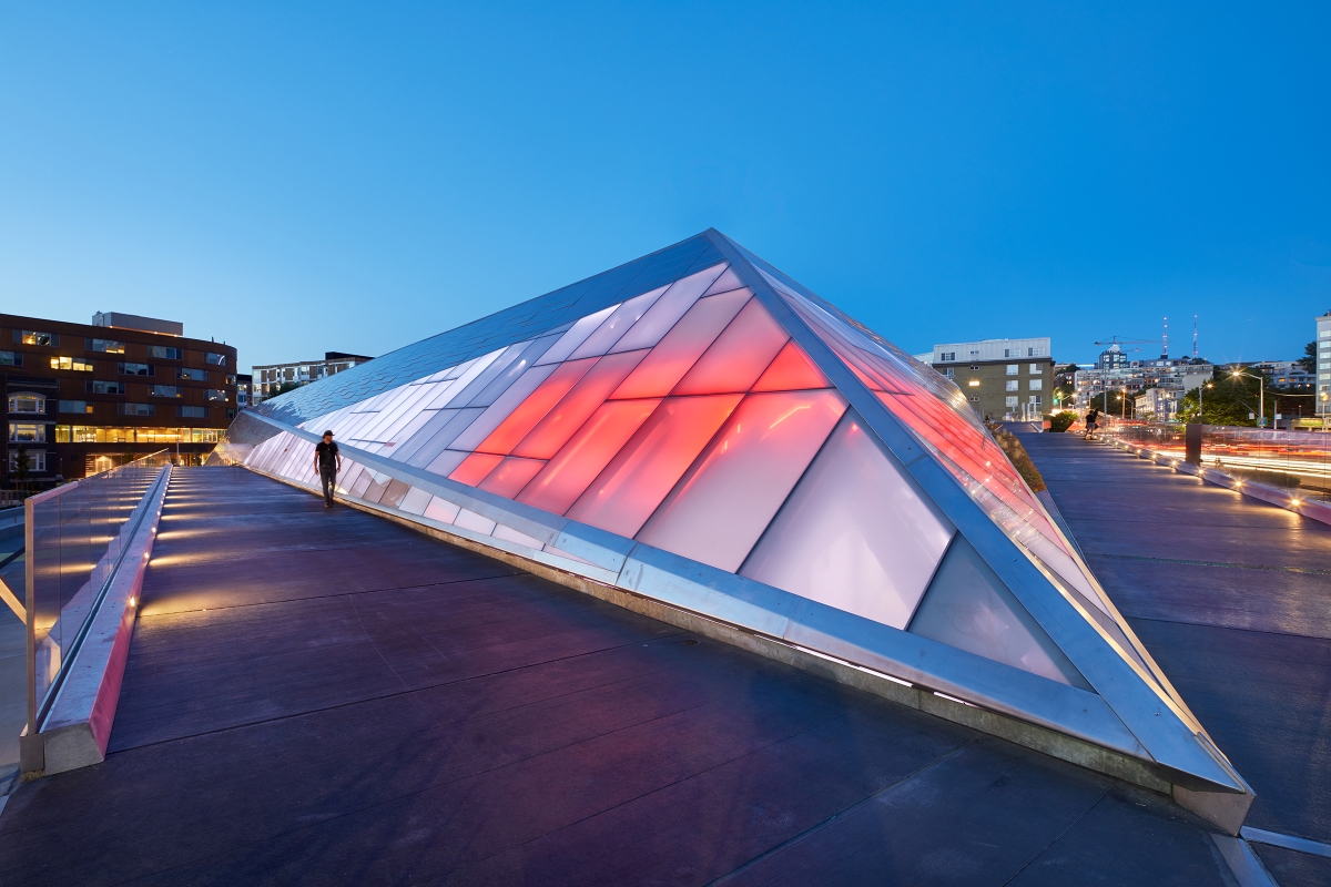 An architectural photograph capturing the modern Denny Substation structure with a geometric, triangular design during twilight. The building features a series of triangular panels, some of which are illuminated from within, casting a warm glow. The structure is surrounded by railings and is part of a larger complex, with other buildings visible in the background under a clear blue sky transitioning to sunset hues.
