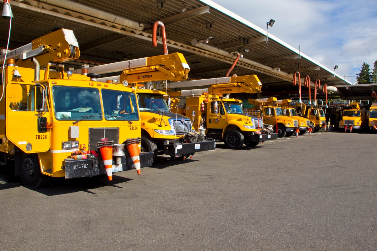 A fleet of yellow Seattle City Light utility trucks with cherry picker booms parked in a row under a clear blue sky. The vehicles are equipped with various compartments and tools, indicating they are service trucks possibly for electrical maintenance. Each truck has reflective stripes and is marked with identification numbers on the side. The setting is a storage or staging area for a utility company, ready for deployment.