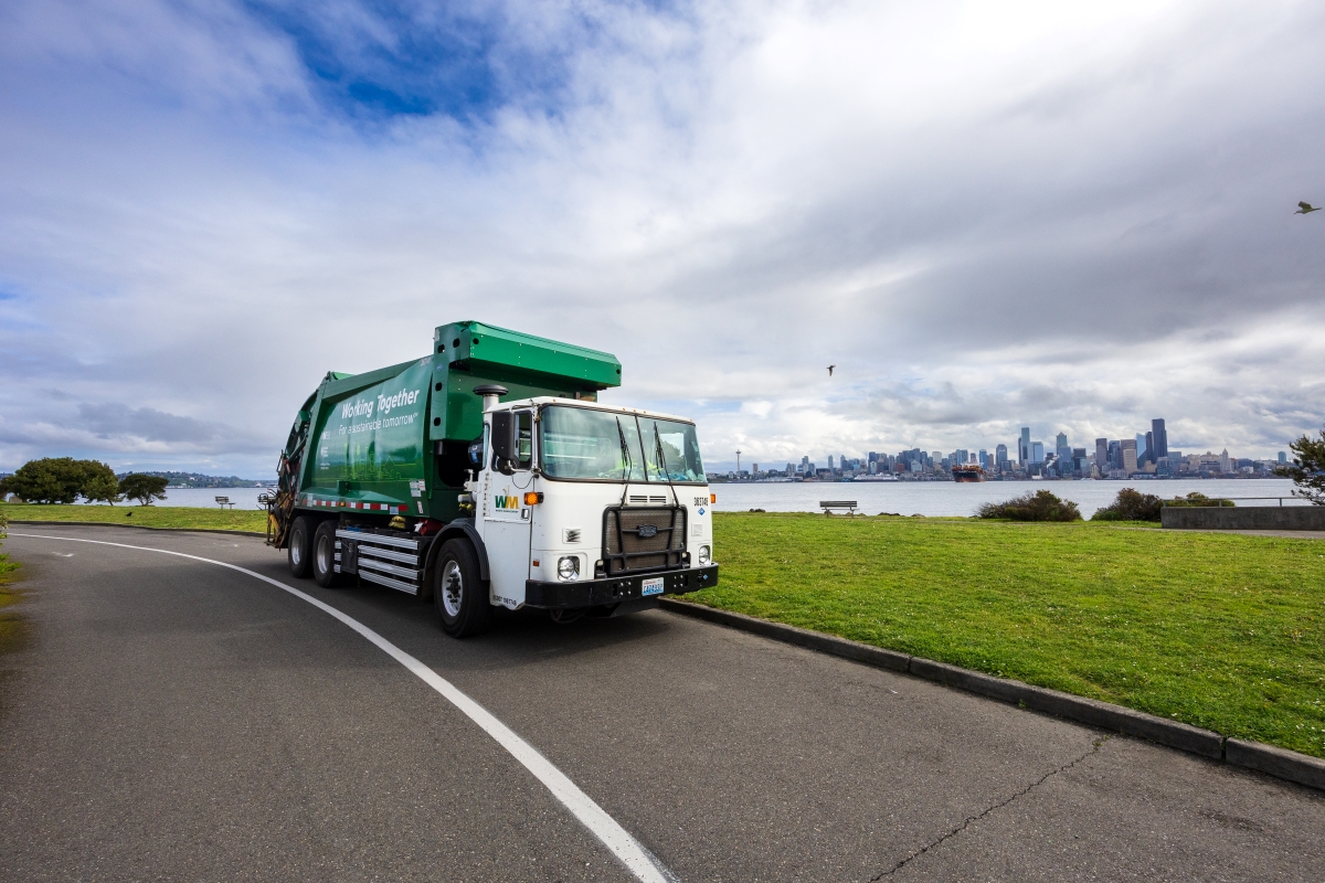 A green and white Seattle Public Utilities waste management truck driving on a paved road with a grassy area to the right along Alki beach. In the background, there is a city skyline under a cloudy sky.