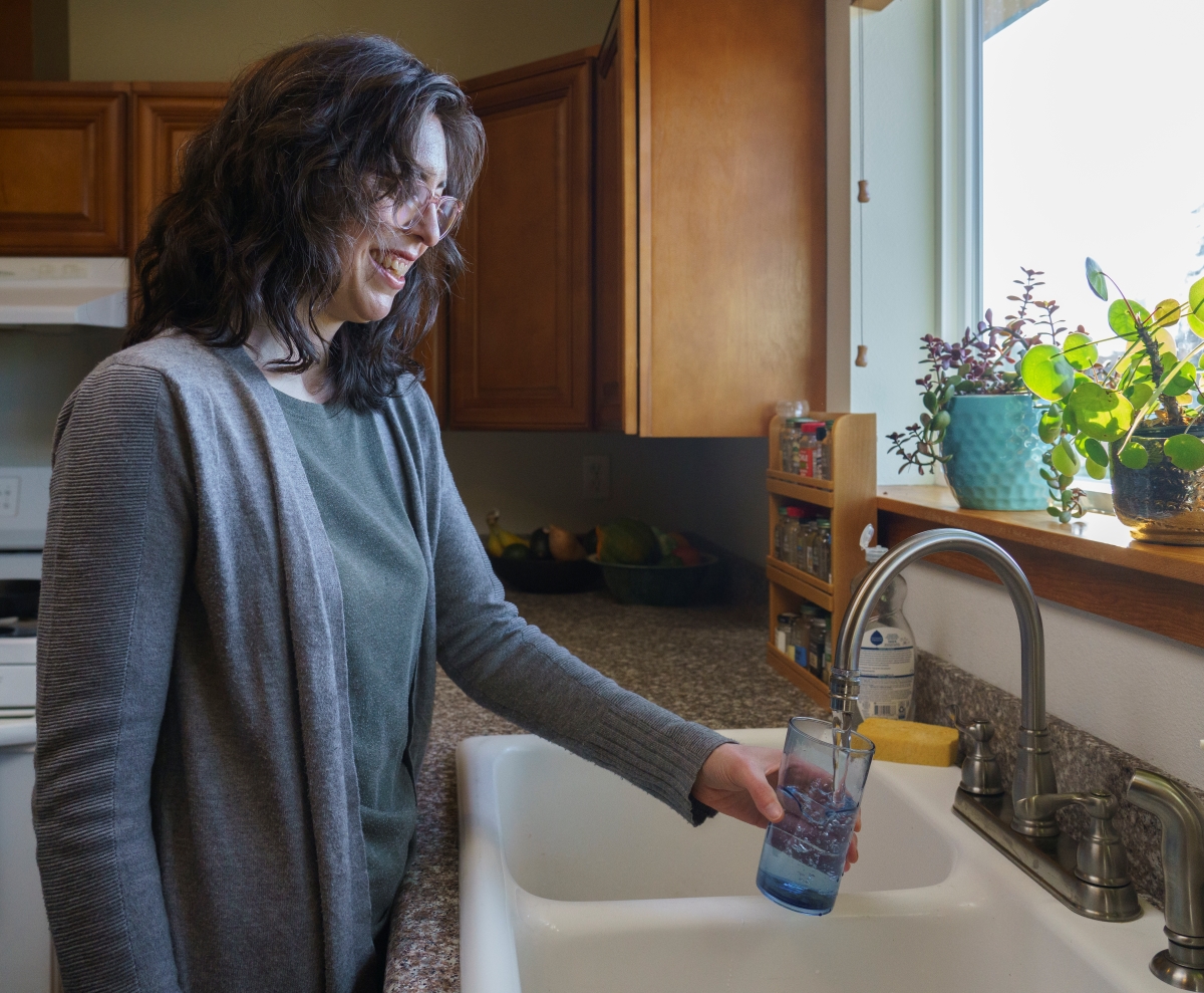 A woman stands at a kitchen sink, filling a clear glass with water from a stainless steel faucet. The kitchen has wooden cabinets and a window through which sunlight is coming in, illuminating some houseplants on the windowsill. There is a bowl of fruits visible on the countertop to the left.