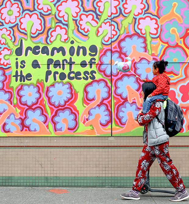 Woman with child on shoulders walks by graffiti mural which says "dreaming is a part of the process," mural by @barelyawakekalee