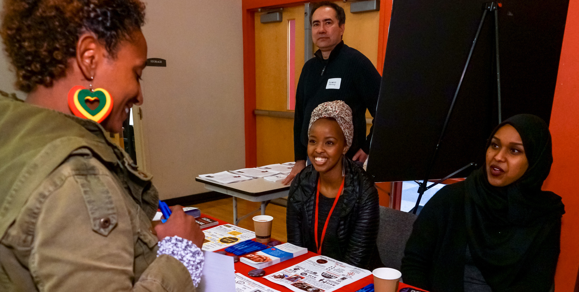 Two young Black women, one wearing a head scarf and the other wearing a hijab are sitting behind a table inside of a room talking to a Black community member who is smiling as she looks at the resources available on top of the table.