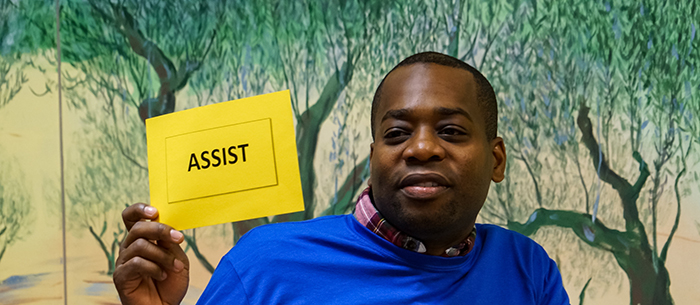 A Black man wearing a bright blue shirt acting as a volunteer at a City of Seattle Citizenship Clinic holds up a small bright yellow sign that reads, "ASSIST".