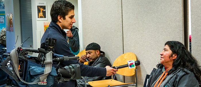 Univision Reporter Pablo Gaviria stands next to a large video camera. He is holding a microphone towards a Latina woman wearing a coat as he interviews her about her experience at a City of Seattle Citizenship Clinic.