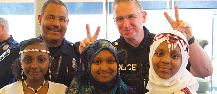 Three East African young women dressed in traditional outfits smiling with two uniformed Seattle police officers standing behind them and also smiling. One of the police officers is making the “bunny ears” gesture with his hands and holding each hand up to behind the heads of two of the East African women.