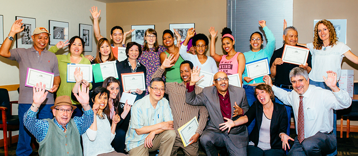 A multiracial group of Ready to Work instructors, administrators, and graduates who are proudly holding up their certificates of completion. Everyone has their hands up in a cheer inside the warmly lit room.