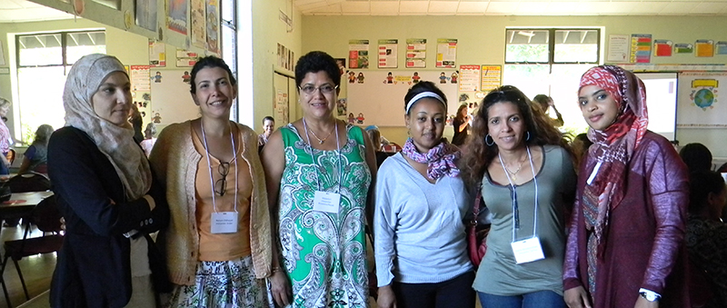 A multiracial group of Refugee Women's Institute members are standing in a line facing you and smiling. Two of the women are wearing hijabs.