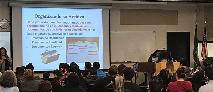 Immigrant attendees at a community event learning about changes to the DACA program from an advocate speaking in front of a slideshow. The slide on the screen features the title: 'Organizando su Archivo.'