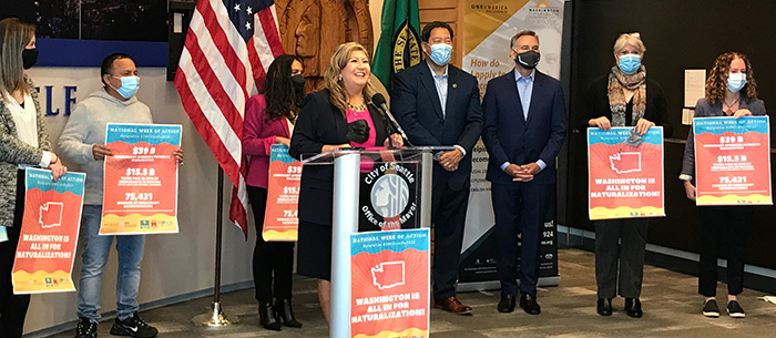 A multi-racial group of people holding signs supporting U.S. citizenship lined up in a row behind a podium.