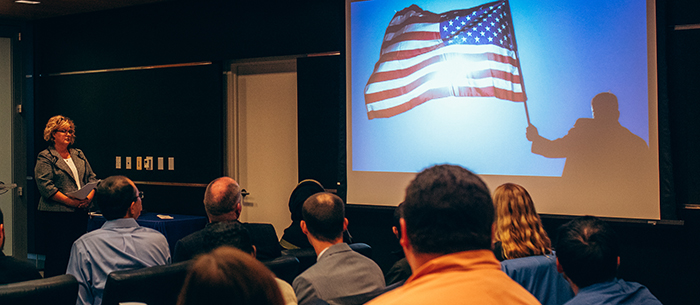 New Americans watching a video about U.S. citizenship in a Seattle City Hall room right before they pledge the Oath of Allegiance. The video features someone waving an American flag.