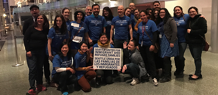 A multiracial group of smiling staff and volunteers most of whom are wearing blue shirts, posing as a group inside the dimly lit McCaw Hall with someone in the center of the group holding a sign that reads: SEATTLE UNITED FOR IMMIGRANT AND REFUGEE FAMILIES | SEATTLE UNIDA POR LAS FAMILIAS DE LOS INMIGRANTES Y REFUGIADOS.