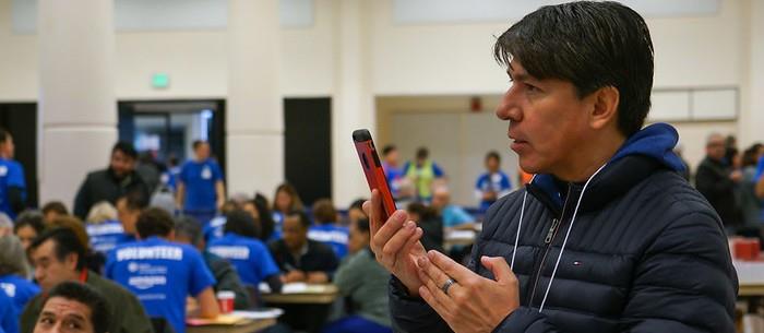 A reporter speaks into his cell phone standing in a school gym with lots of people in the background sitting at desks talking with service providers.