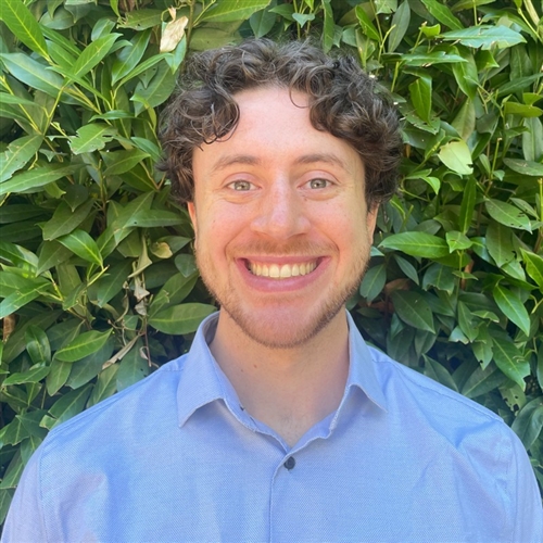 Portrait of Matthew Malloy smiling broadly in a blue collared shirt in front of green foliage.