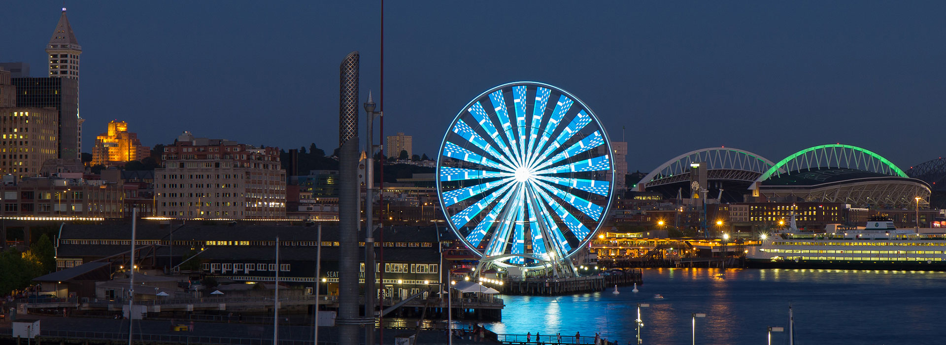 Nighttime waterfront scene featuring a brightly lit Ferris wheel, surrounding city buildings, and a stadium with illuminated arches.
