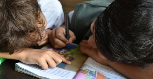two children looking at a workbook. one child writes while the other points to the book