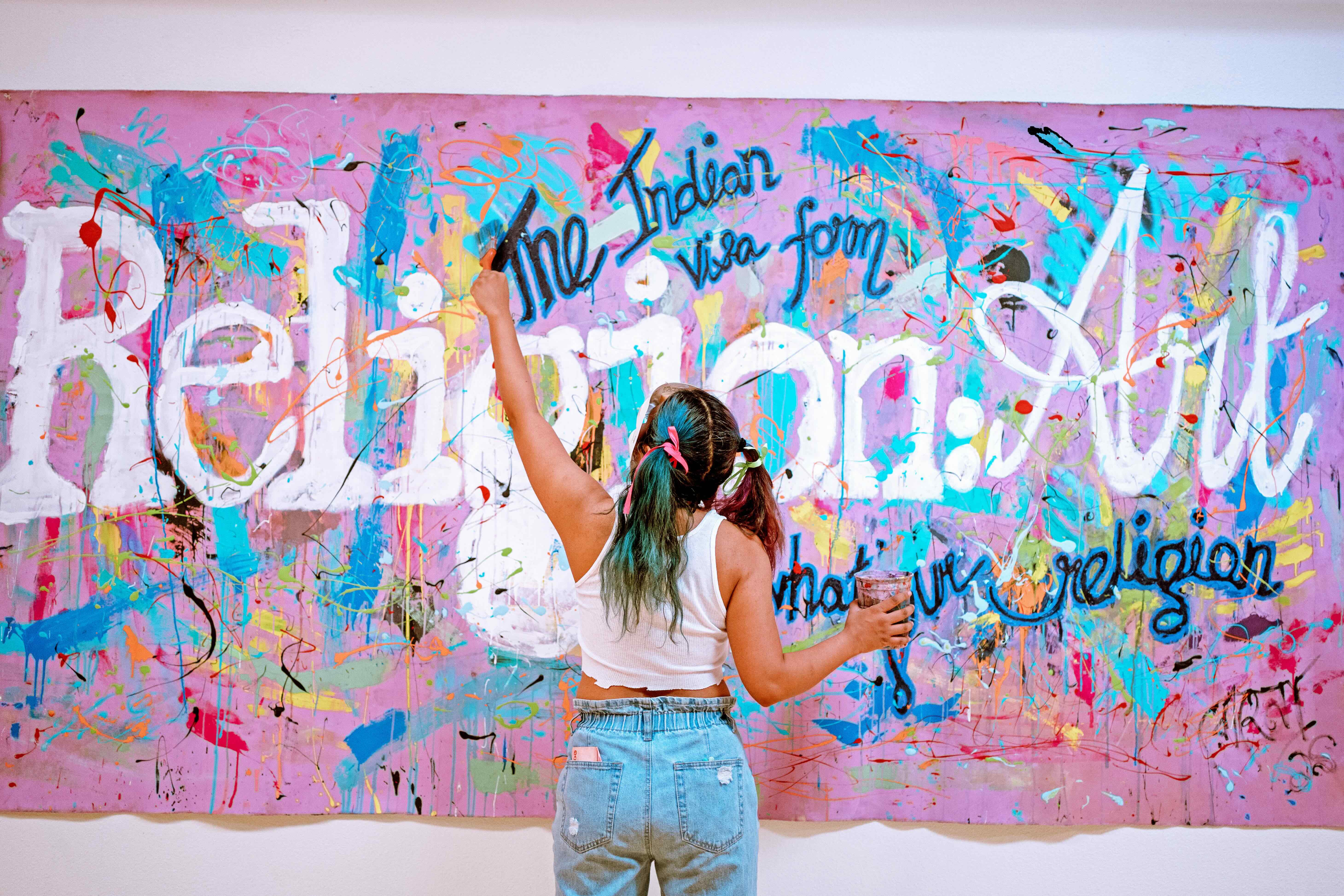 Rohena has blue hair and two pigtails. She reaches high to paint on a large colorful canvas with paint splatters and text that reads "The Indian Visa Form, What's your religion; Religion:Art"