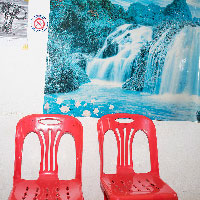 Two plastic red chairs against the wall. A torn, lopsided and faded poster of a waterfall hangs behind along with a No Smoking sticker.