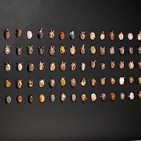 Many small clay masks of different designs and glazes displayed on a black wall.