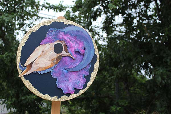 A circular collage of a goat skull mounted on a wooden stick displayed in front of trees.
