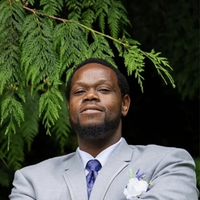 Joshua has dark skin, close cropped hair, a beard, and smiles in a grey suit.