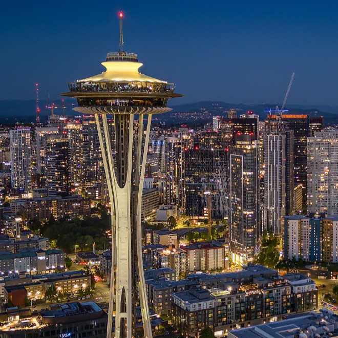 An ariel view of the Seattle skyline at night with building lights lit up, the Space Needle is featured prominently in the shot.