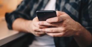 A person in a grey and black plaid flannel button down shirt with a white shirt underneath is holding a mobile phone device in both hands. The focus of the image is on the mobile device.