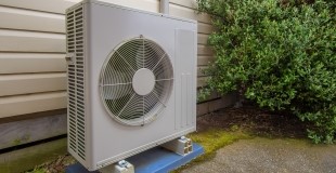 An outdoor unit of a split-system air conditioner placed on a concrete pad beside a house with vinyl siding. The unit has a large circular fan in the center and is surrounded by green shrubbery in a residential setting.