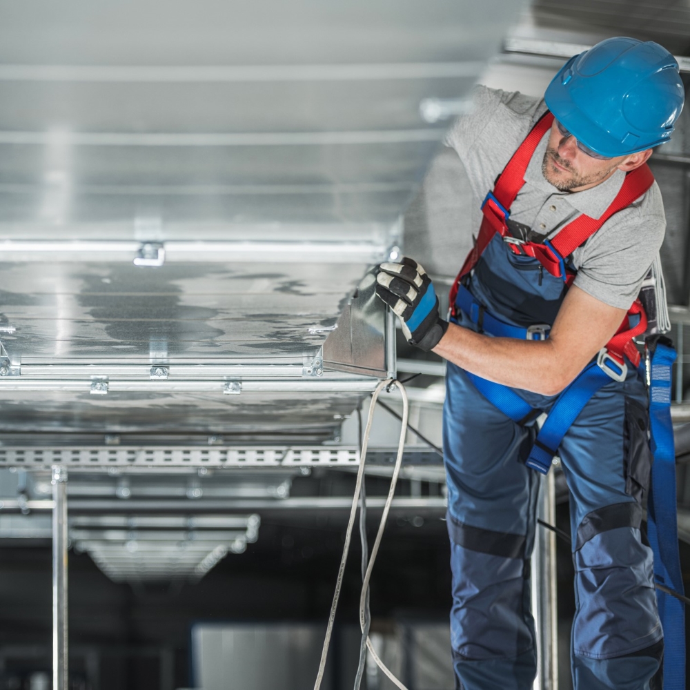 A person wearing a safety harness is working on an air duct in a commercial building. The individual is using a drill to secure or adjust the ductwork. The setting is maintenance work being performed in an industrial or commercial environment.