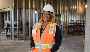 Image depicts African American preschool director standing at new facility construction site and wearing a hard hat and safety vest.
