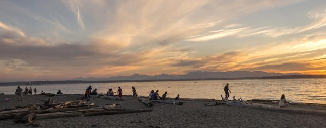 Image of Golden Gardens beach with people on the beach enjoying a golden sunset. 