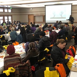 Crowded room of attendees seated at table listening to a presenter in front of a screen