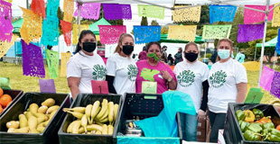 Group of women standing behind a produce table with colorful flags in the background