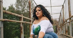 Roxanna Pardo Garcia crouches on a bridge looking serious with long curly hair and spray-painted t-shirt. 