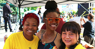 Group of three smiling women at a festival under a tent.