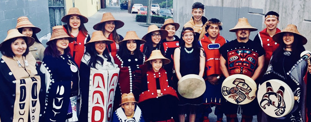 Group of Haida in traditional dress wearing hats and holding handmade drums with beautiful Haida imagery.