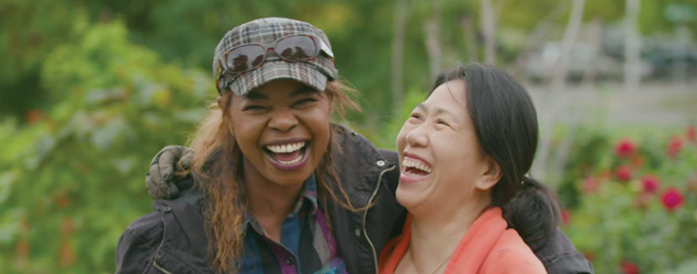 Two women side-hugging and laughing in a p-patch garden laden with red poppy blossoms and lush foliage