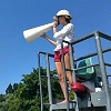 A lifeguard shouts through megaphone atop a lifeguard chair, somewhere outdoors.