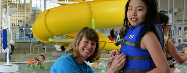 Adult woman putting a life vest on a child; both are smiling. There is a twisty, yellow slide into the pool behind them.