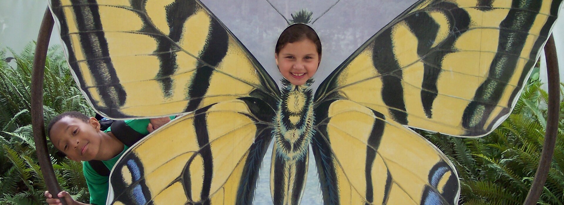 Little girl poking her head through a large yellow butterfly cutout, a little boy peeks around the side