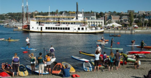 Kayakers and paddleboarders depart from the beach of Lake Union Park. In the background, a ship is docked at the wharf.